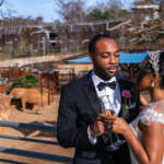 On a balcony overlooking the elephant habitat, a couple dressed in formal attire pose while toasting glasses champagne.