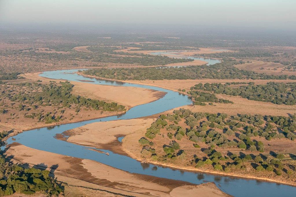 A river twists through a brown landscape accented by groups of green trees.